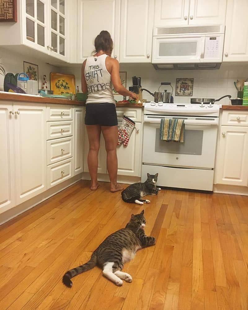 A picture of Donna making food in the kitchen with two cats lying on the floor near her feet.
