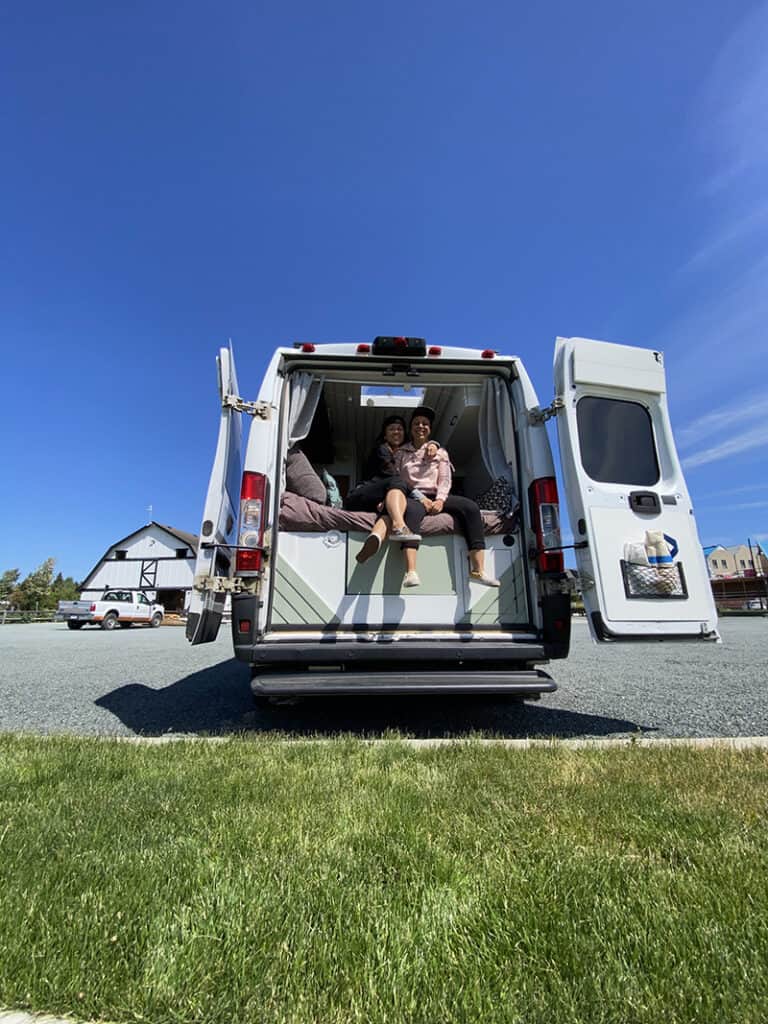 A picture of our campervan from the back with the doors open and the two of us sitting on the bed.